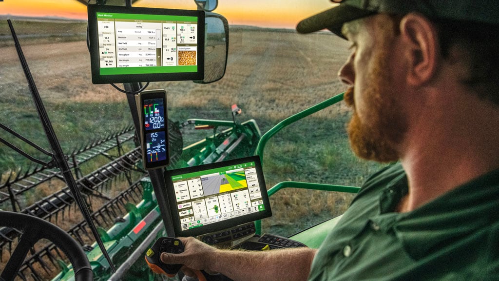 Photo en cabine d’un agriculteur conduisant une moissonneuse-batteuse John Deere dans un champ au coucher du soleil. Il regarde une console G5Plus allumée. Il dispose également d’un moniteur étendu G5Plus au-dessus d’une console d’angle principale.