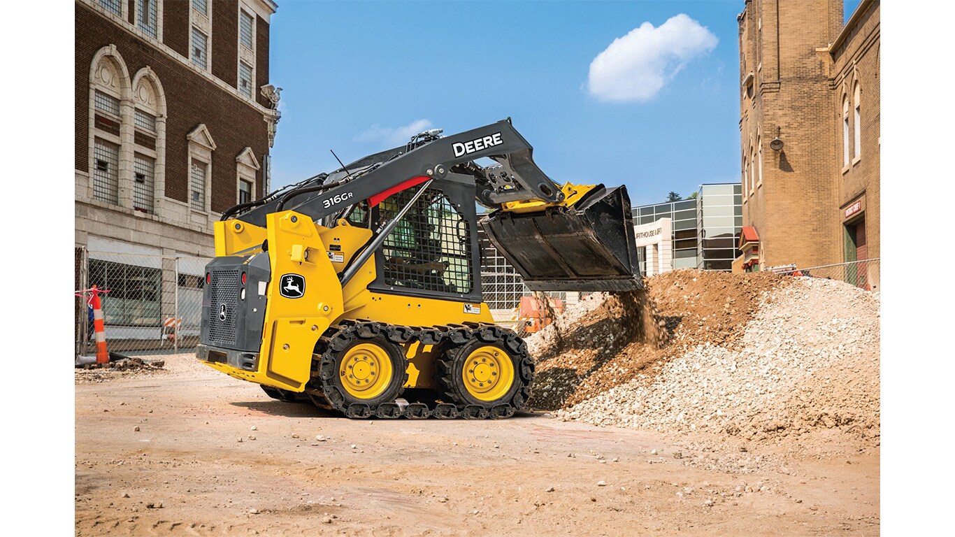 Une chargeuse à direction différentielle 316G déverse des roches et de la terre dans une pile sur un chantier avec des bâtiments en arrière-plan.