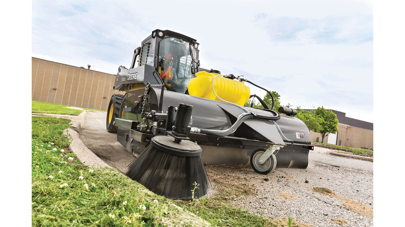 A 320G Skid Steer with a pick-up broom attachment sweeps grass clippings off the road and gutter.
