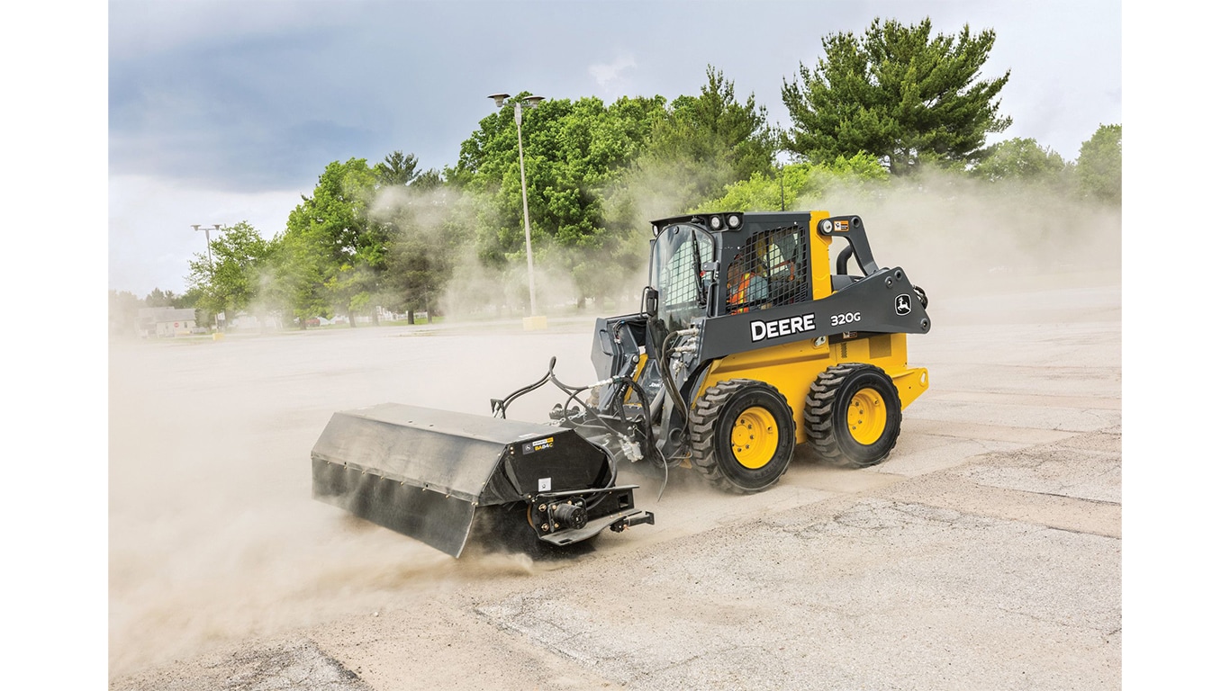 A 320G Skid Steer with pick-up broom attachment sweeps dirt and dust off a parking lot.