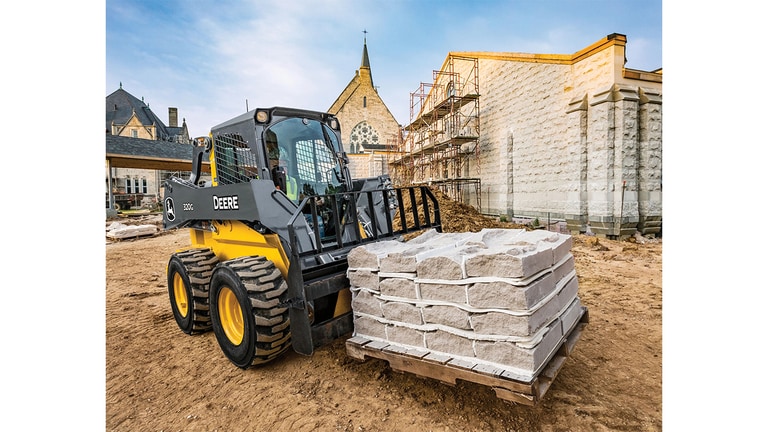 A 320G Skid Steer with pallet attachment transporting a pallet of stones with a church in the background.