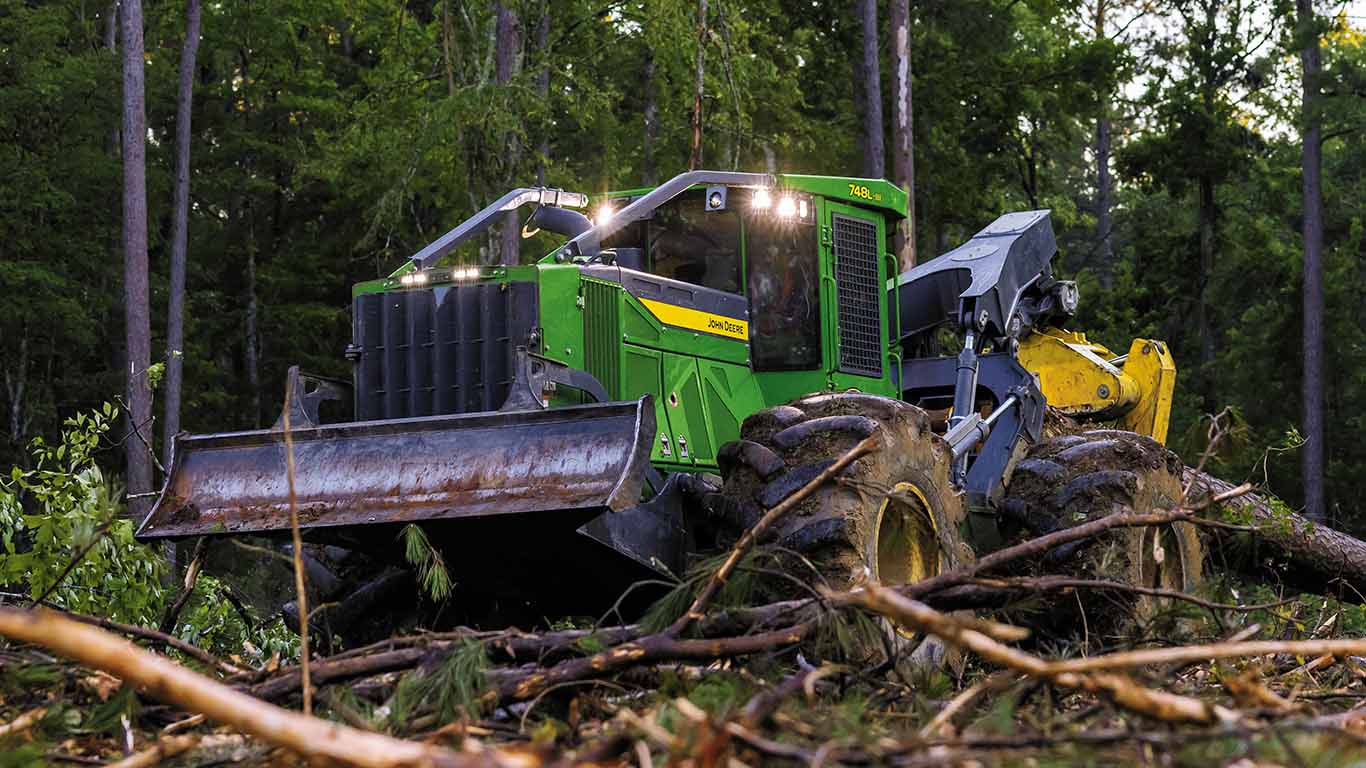 Une&nbsp;débardeuse verte dotée d'une lame et d'une pince ramasse des grumes dans une forêt dense.