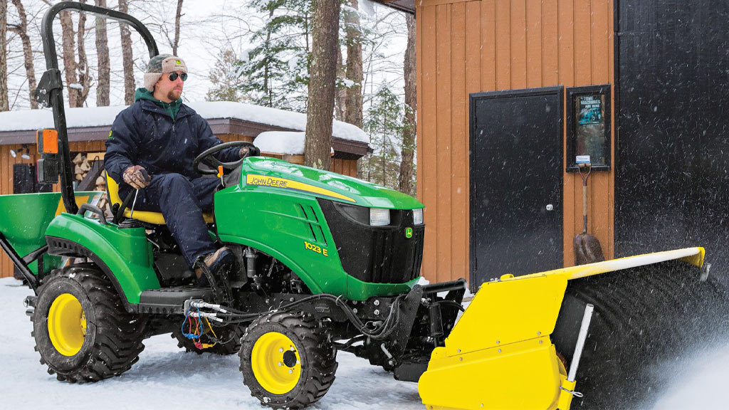 Un homme conduit un tracteur compact 1023E déblayant la neige avec un balai rotatif
