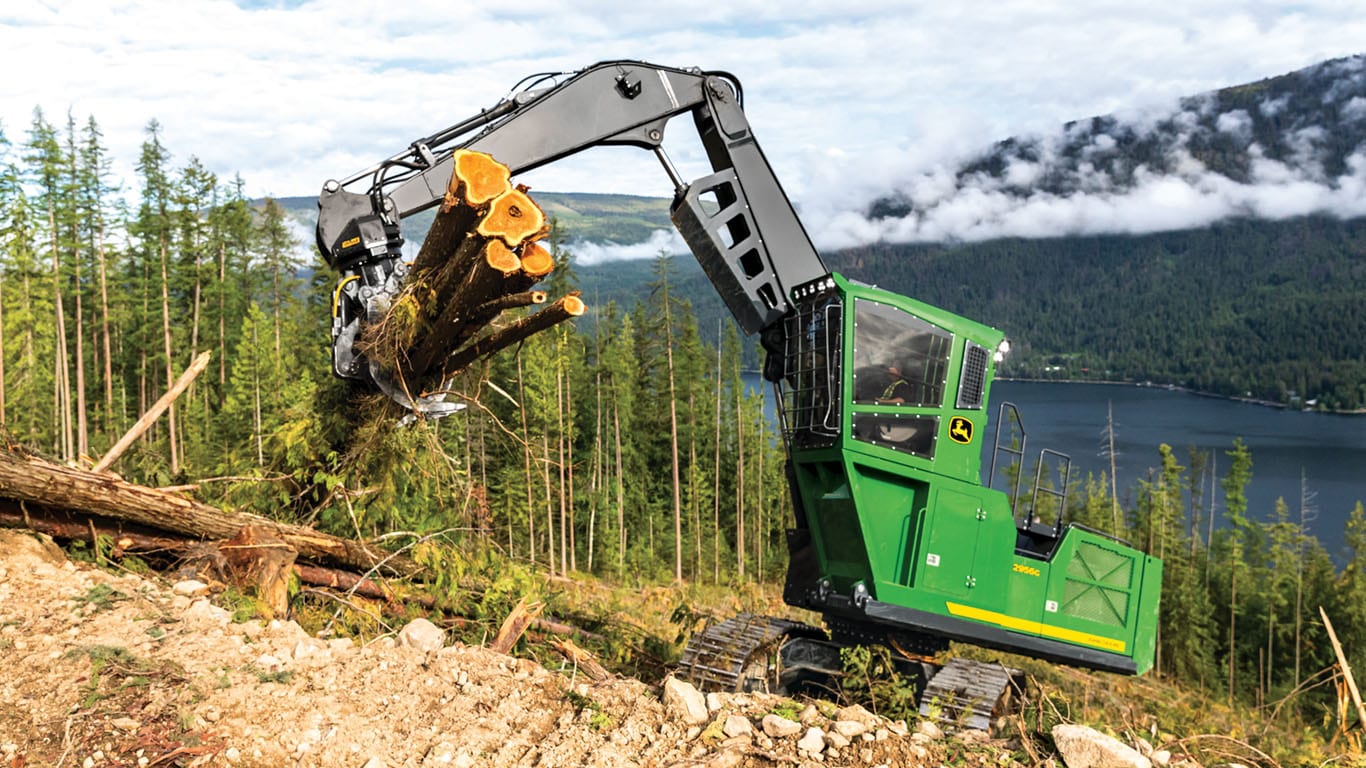 Un engin pivotant de John&nbsp;Deere soulevant des rondins avec un lac et des arbres en arrière-plan.