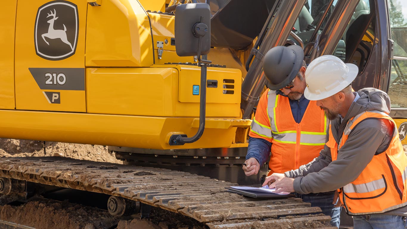 Deux ouvriers de la construction portant des équipements de sécurité examinent des documents sur une planchette à pince à côté d’une excavatrice jaune.