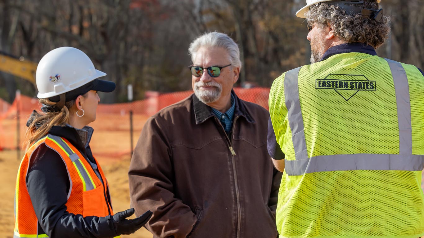 Trois personnes portant de l’équipement de sécurité discutent sur un chantier de construction entouré de clôtures oranges, avec des arbres en arrière-plan.