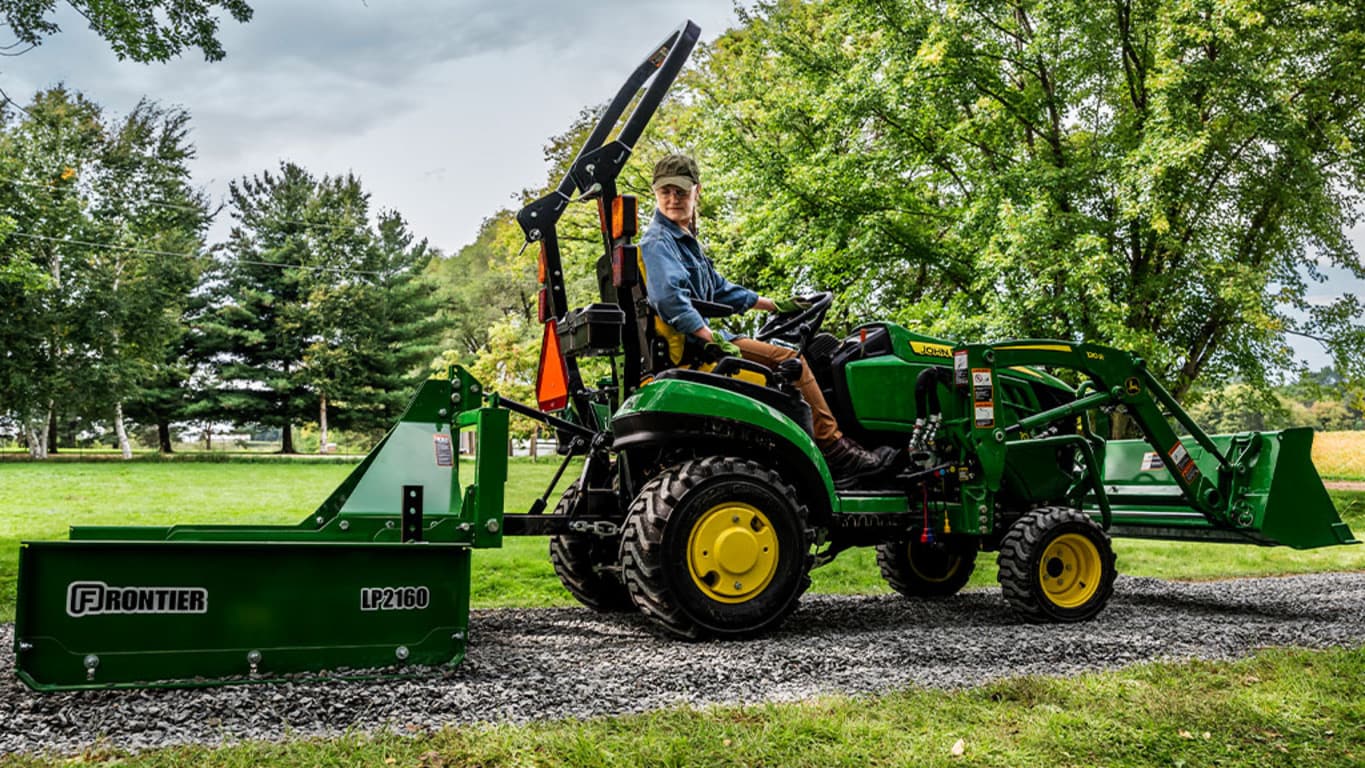 A person looks back as they level gravel with a land plane pulled by a green compact tractor.