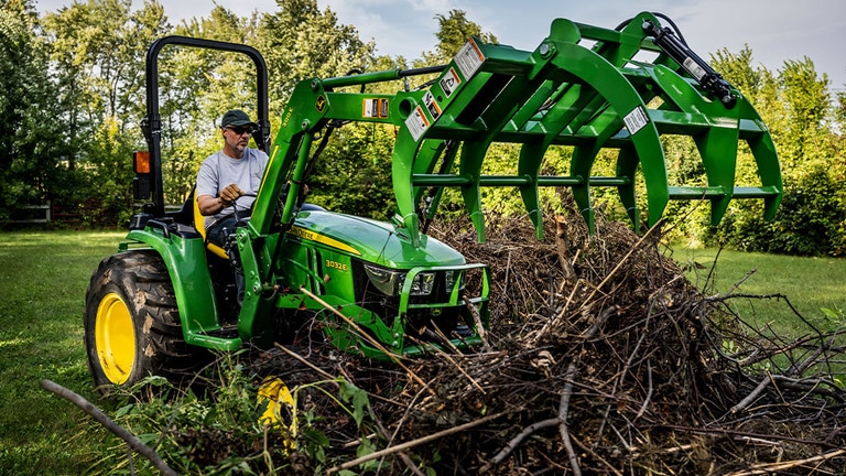 Un grappin à débris fixé à un tracteur compact vert est ouvert, prêt à saisir un tas de branches