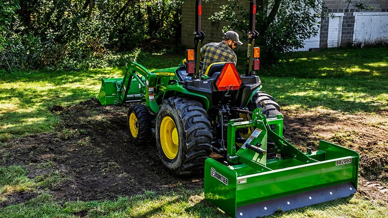 Un chargeur frontal et une boîte niveleuse montés sur un tracteur compact vert raclent la couche de terre superficielle et l'herbe pour créer une surface de terre.