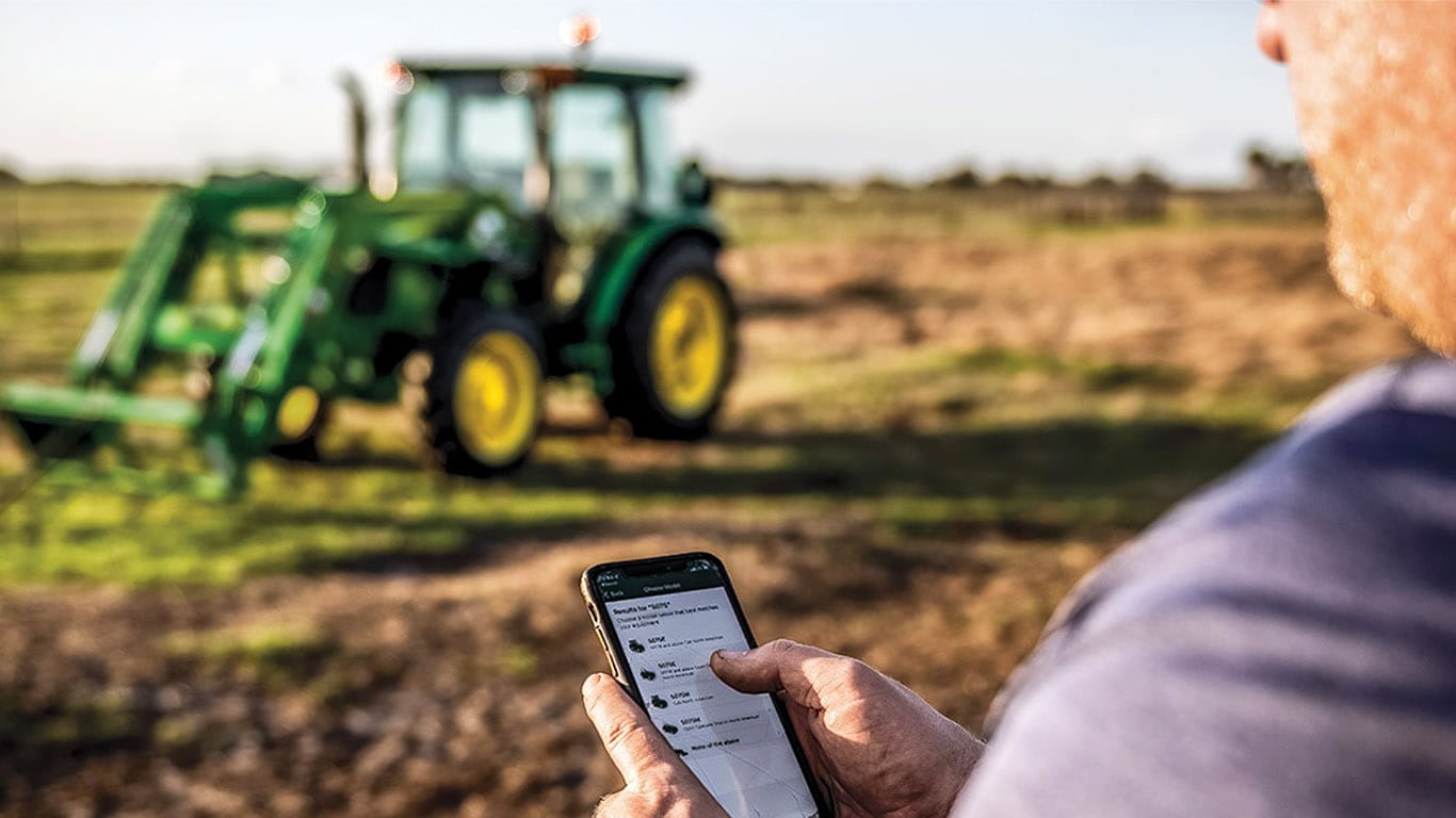 Personne utilisant Property Center™ sur un appareil mobile avec un tracteur de la série 5 dans le champ, en arrière plan.