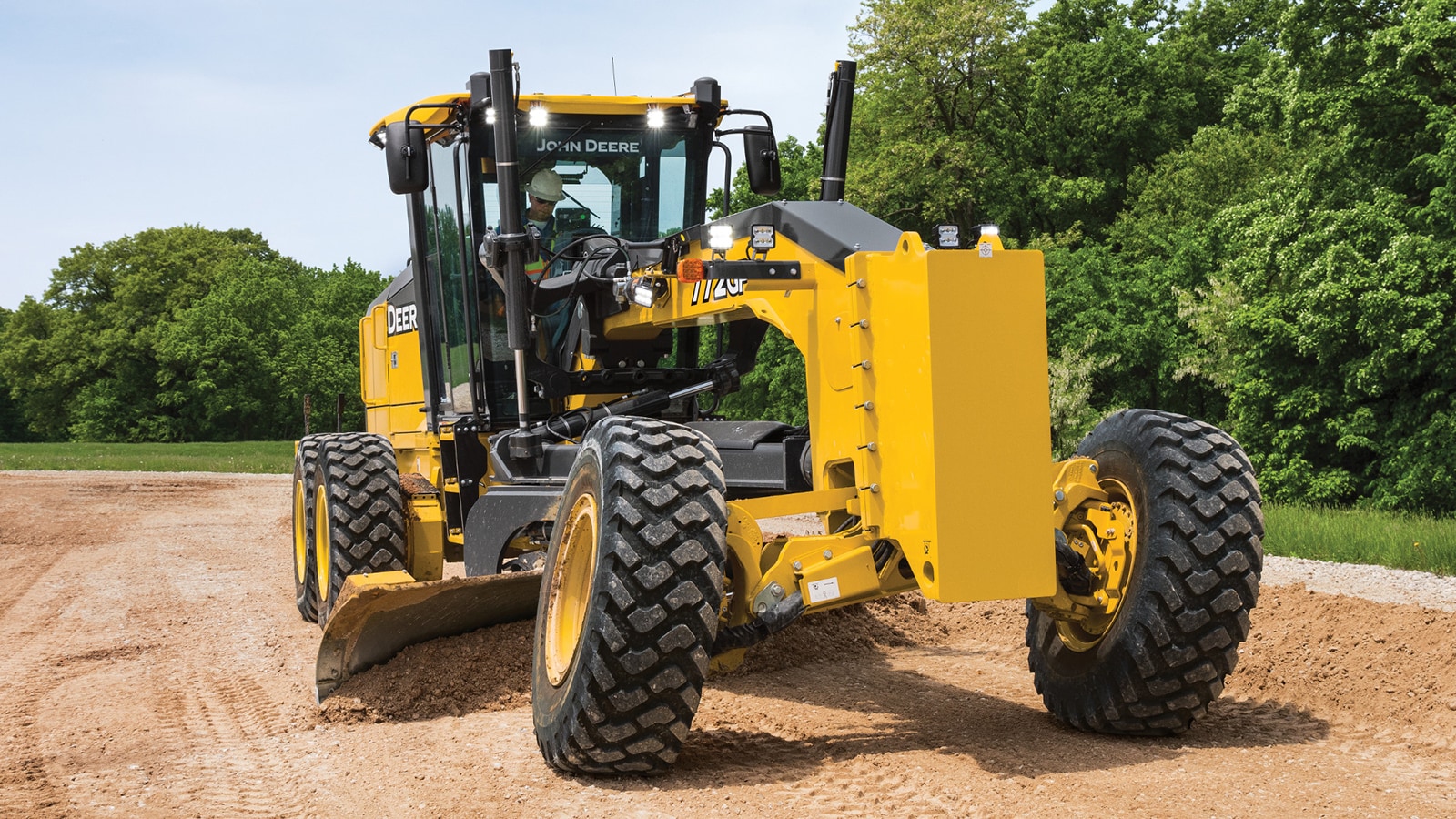 John Deere 772GP SmartGrade Motor Grader grades a dirt lot surrounded by trees