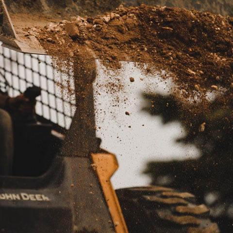 A John Deere skid steer, tilting its bucket to dump out dirt.