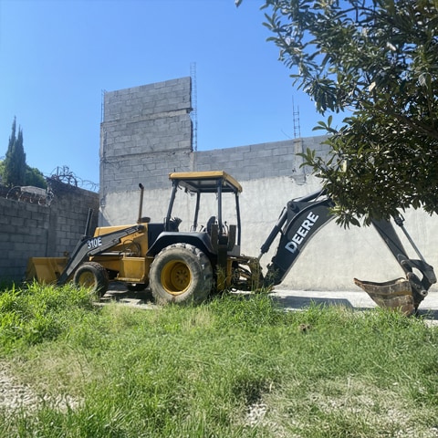 A John Deere 310E backhoe loader sits in front of cinder block walls with with barbed with sitting on top.