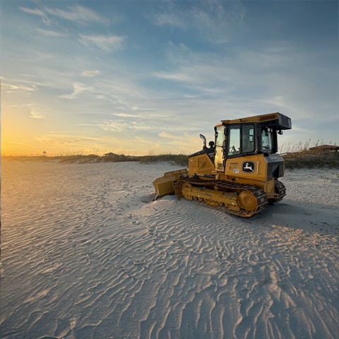 A John Deere 650 P-Tier Dozer sitting on the beach facing a sunset.