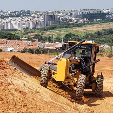 Niveleuse automotrice nivelant une pente avec des maisons en arrière-plan.
