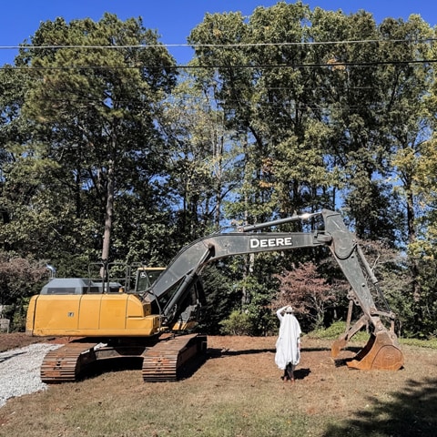 A John Deere 210G LC Excavator sitting in front of trees with a person dressed as a ghost under the boom arm.