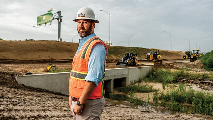 Mike Reinke, vice-président de Prince Contracting, pose sur un chantier de construction routière en Floride alors que des chargeuses compactes chenillées et des bouteurs travaillent en arrière-plan. 