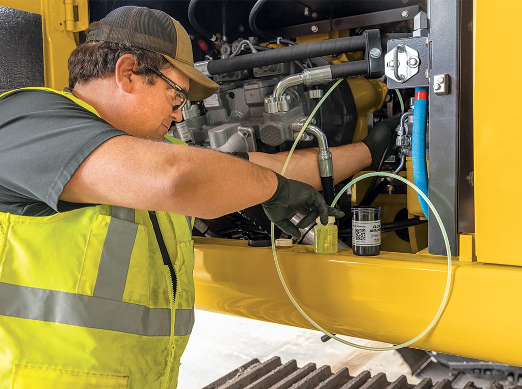 Chat takes a sample of hydraulic oil for a fluid analysis test as he stands beside the open service panels of an excavator.