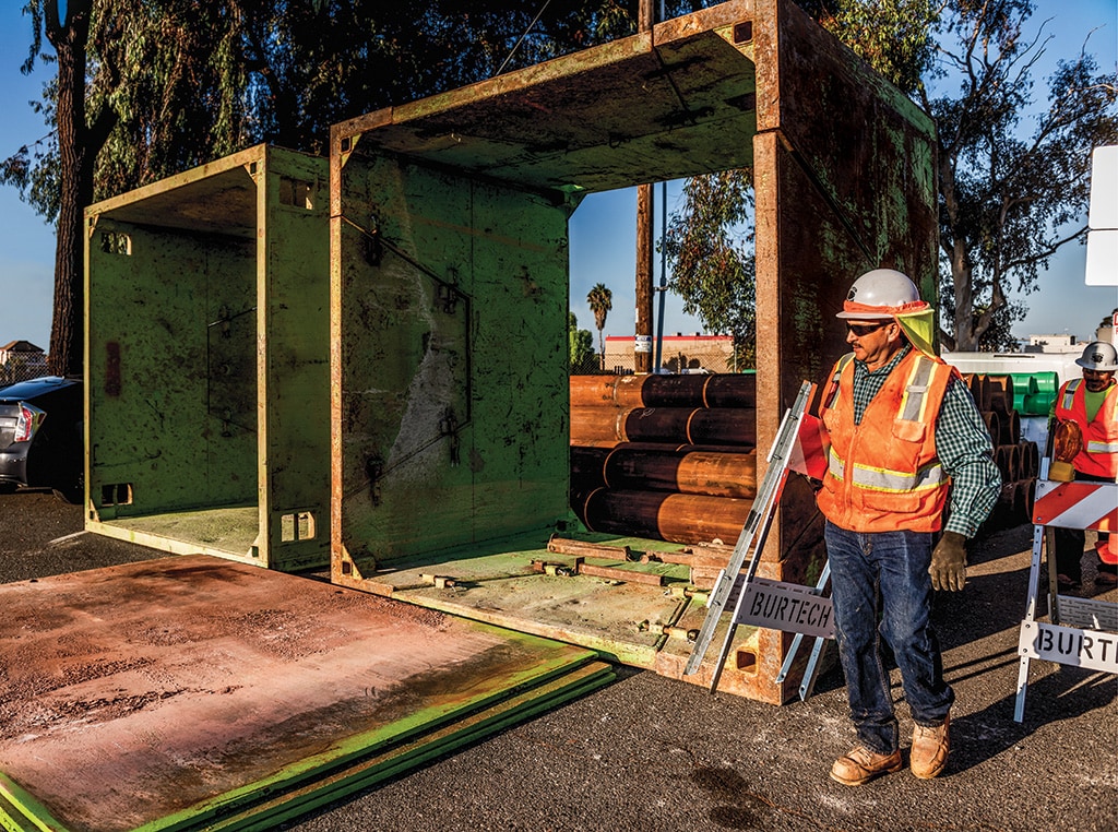 Saul carries a folding traffic barricade as he walks through the jobsite and passes two trench boxes ready for use.