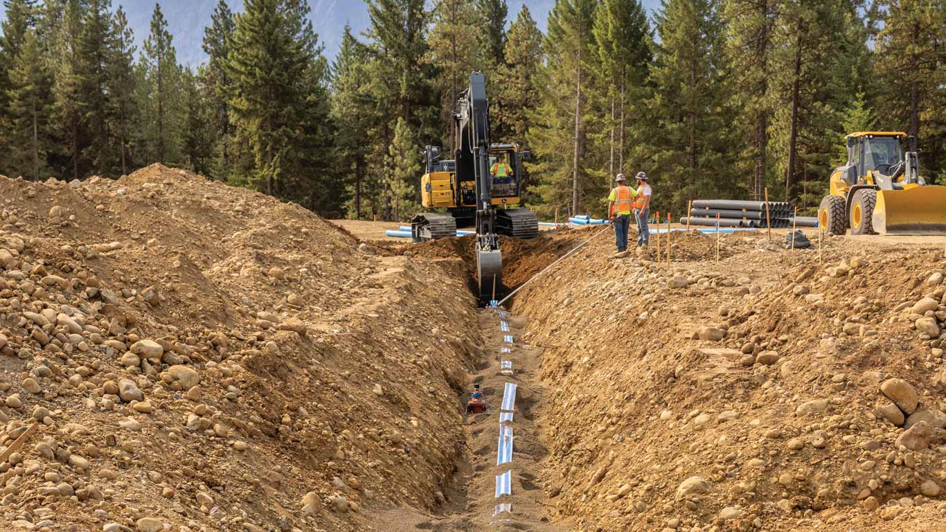 Workers install piping in a trench while an excavator operates at a forested construction site.