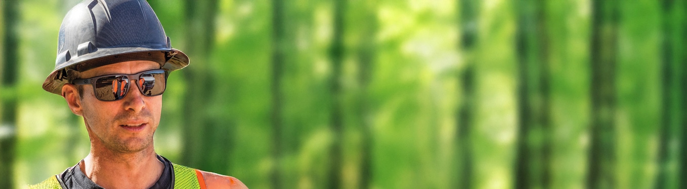 Person wearing a hard hat and safety vest stands outdoors with green trees in background.