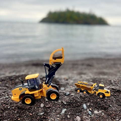 A John Deere 544 P-Tier Wheel Loader and John Deere 460E-II Articulated Dump Truck toy positioned on a dark pebble beach, creating a "sandbox quarry".