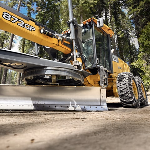 A John Deere 872 GP Motor Grader with chains on its tires with trees in the background.