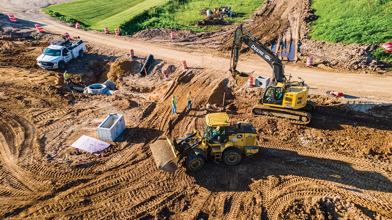 A John Deere 624 P-Tier hauls gravel to an exposed manhole and a 245 G-Tier Excavator fills a trench.