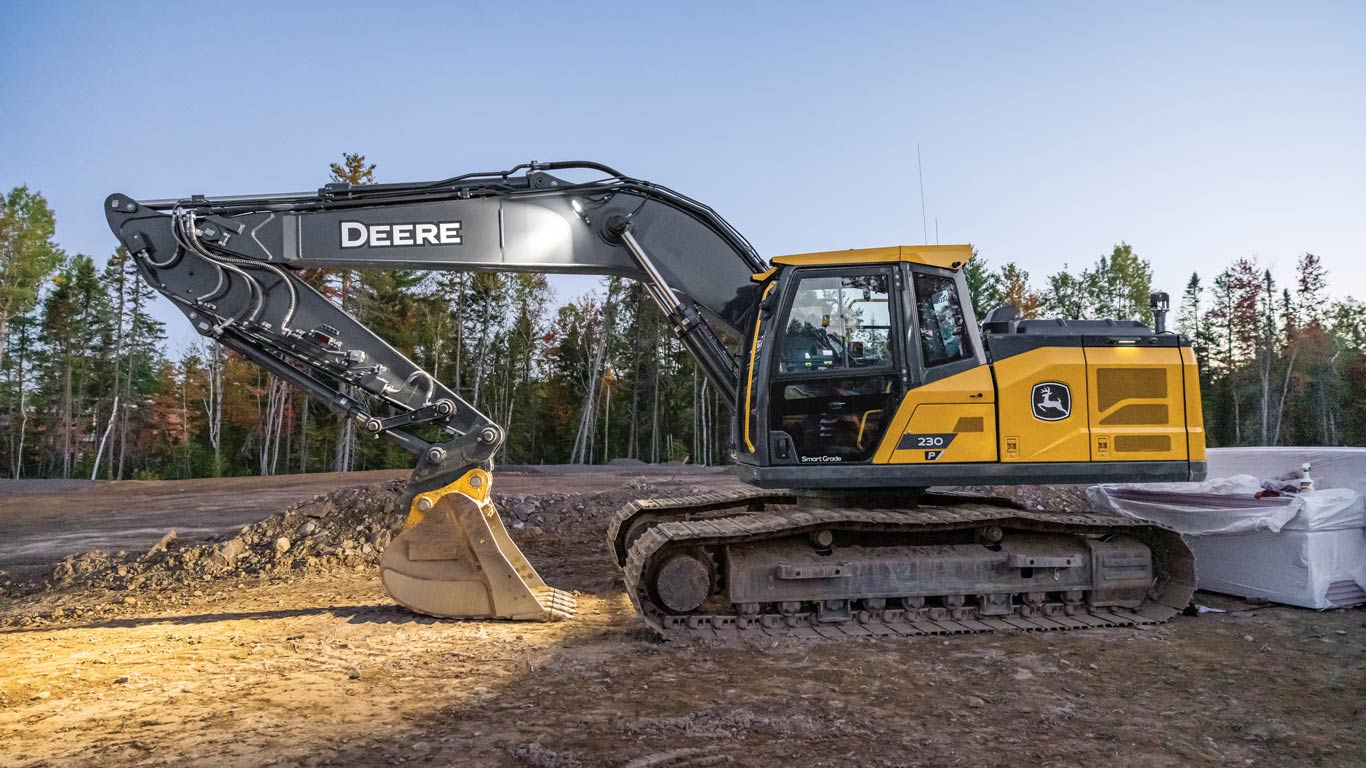 John Deere tracked excavator parked at a construction site with barriers and trees in the background.
