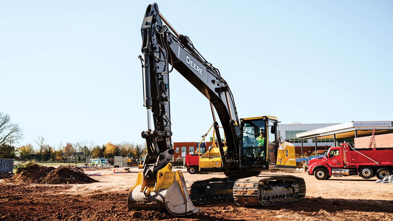 Tracked excavator digging soil at an active construction site with trucks and buildings nearby.