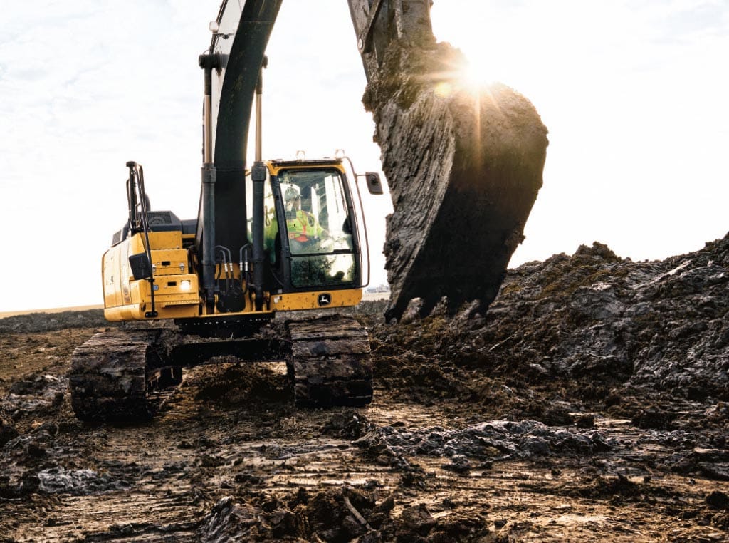 A 210G LC SmartGrade Excavator empties the wet dirt from its bucket next to large piles of dirt.