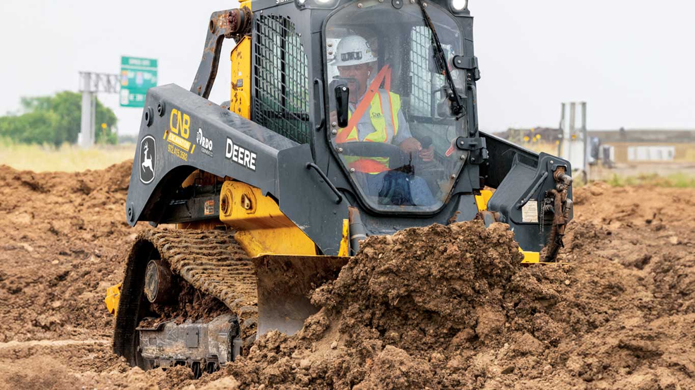 Travailleur portant un équipement de sécurité conduisant une chargeuse John Deere sur un chantier de construction boueux.