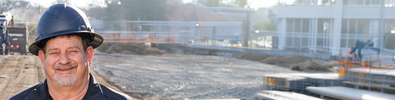 Smiling construction worker with black hardhat on standing in front of a work site