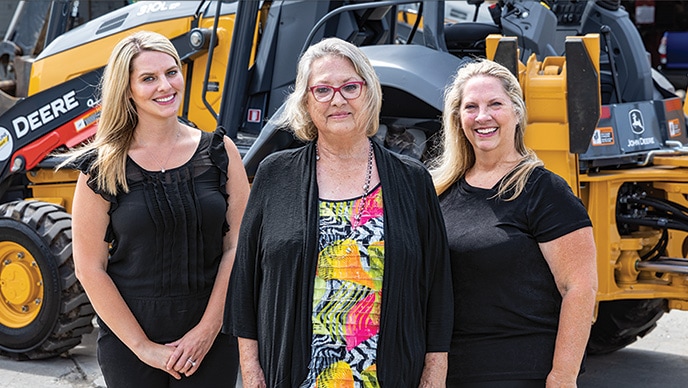 Amanda Matson, Janet Ferguson et Nikki Worden de Newman Backhoe Service se tiennent devant une rétrocaveuse 310L de John Deere.