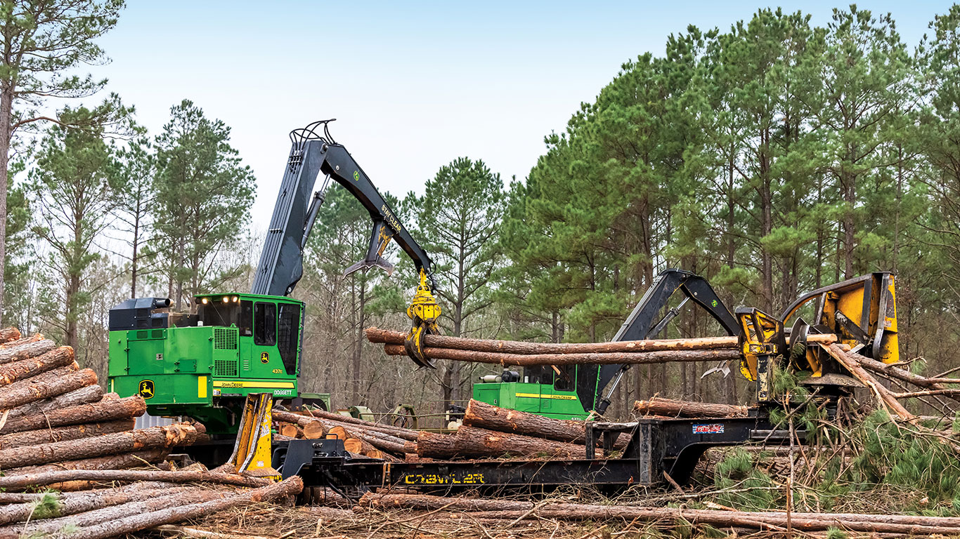 Heavy machinery, including a green John Deere, processes logs in a timber clearing surrounded by tall pine trees.