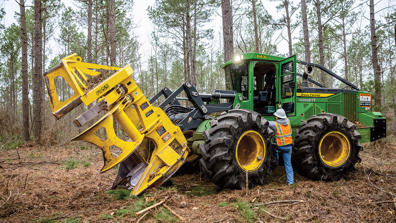 A forestry machine with a yellow attachment operates in a wooded area, while an operator monitors the process from the cab.