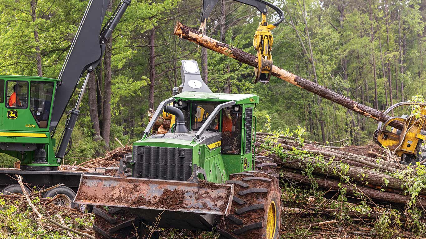 Two green forestry machines in woods; one lifts a log with a grapple.