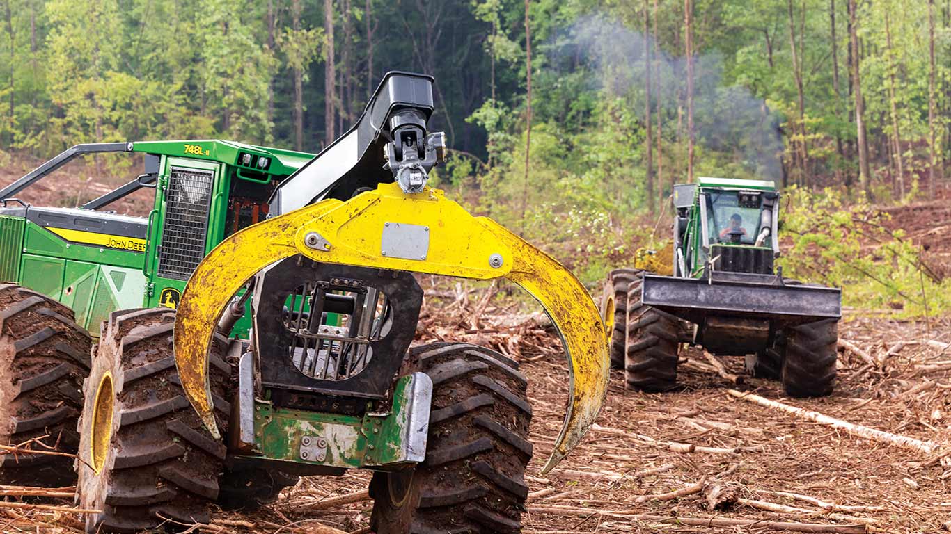 Two skidders in a forest clearing; one lifts logs with a grapple, another emits exhaust.