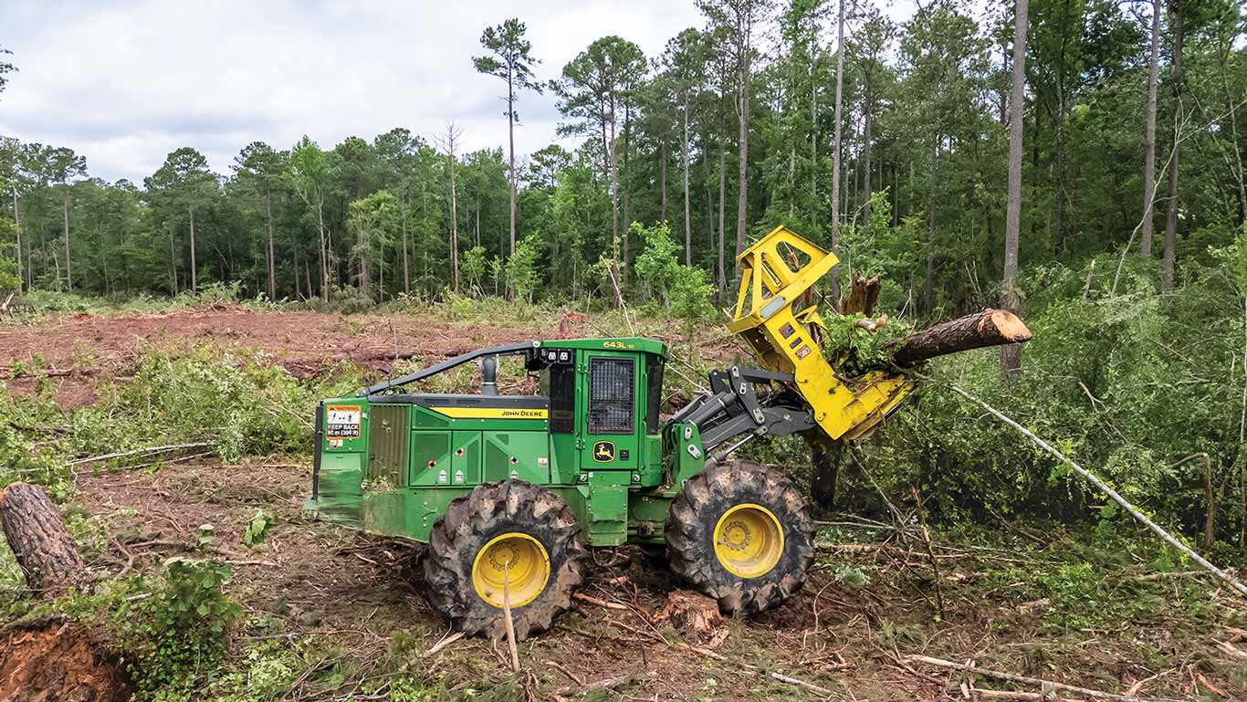 Green skidder with yellow grapple holds log in forest with felled trees and cleared ground.