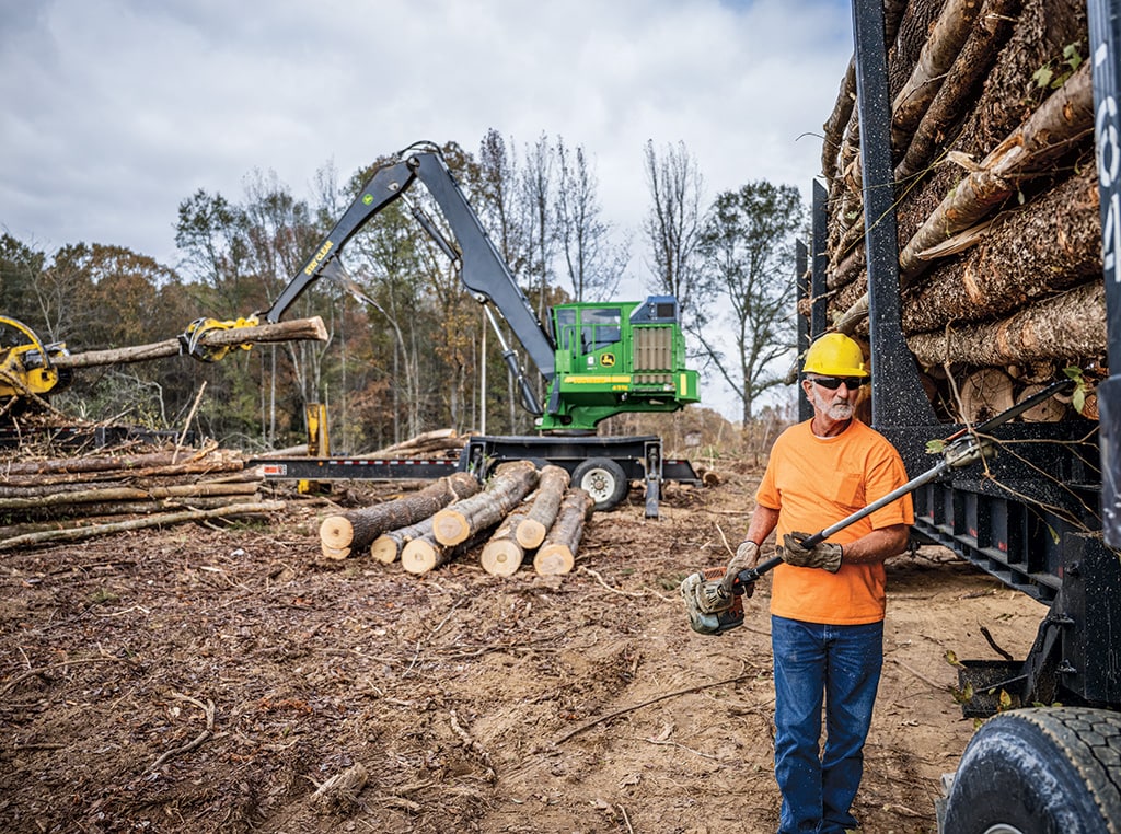 Terry coupe les bûches roulottes alors que la chargeuse à griffe 437E continue de déplacer les arbres coupés.