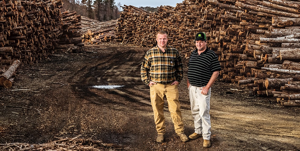 Son Ryan poses to the left of his dad Edwin on the logging road with stacked hardwood on both sides of them at the Edwin Taylor & Son Logging woodyard in Duck Hill, Mississippi.