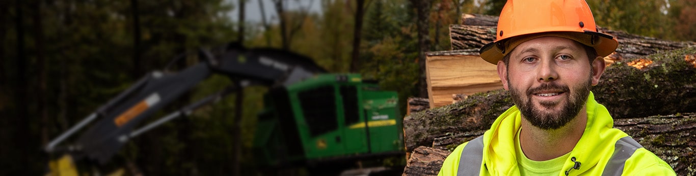 Zachary Emerson, assis sur une pile de rondins et une abatteuse-groupeuse chenillée 853M John Deere avec une tête d’abattage FR22B en arrière-plan.