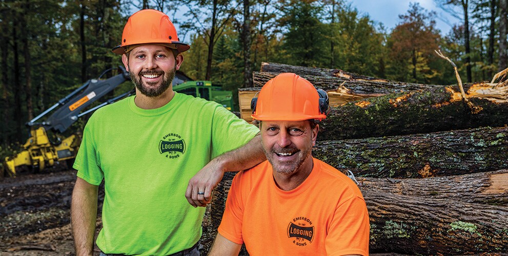 Zachary Emerson, left, and Chris Emerson, right, stand in front of a log pile with a John Deere 853M Tracked Feller Buncher in the background.