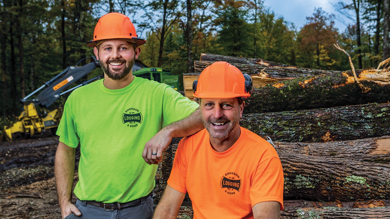 Zachary Emerson, left, and Chris Emerson, right, stand in front of a log pile with a John Deere 853M Tracked Feller Buncher in the background.