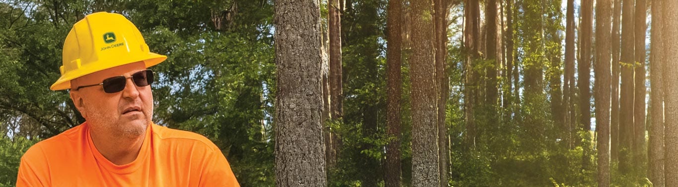 Person in orange shirt and yellow hard hat standing in forest with tall trees.