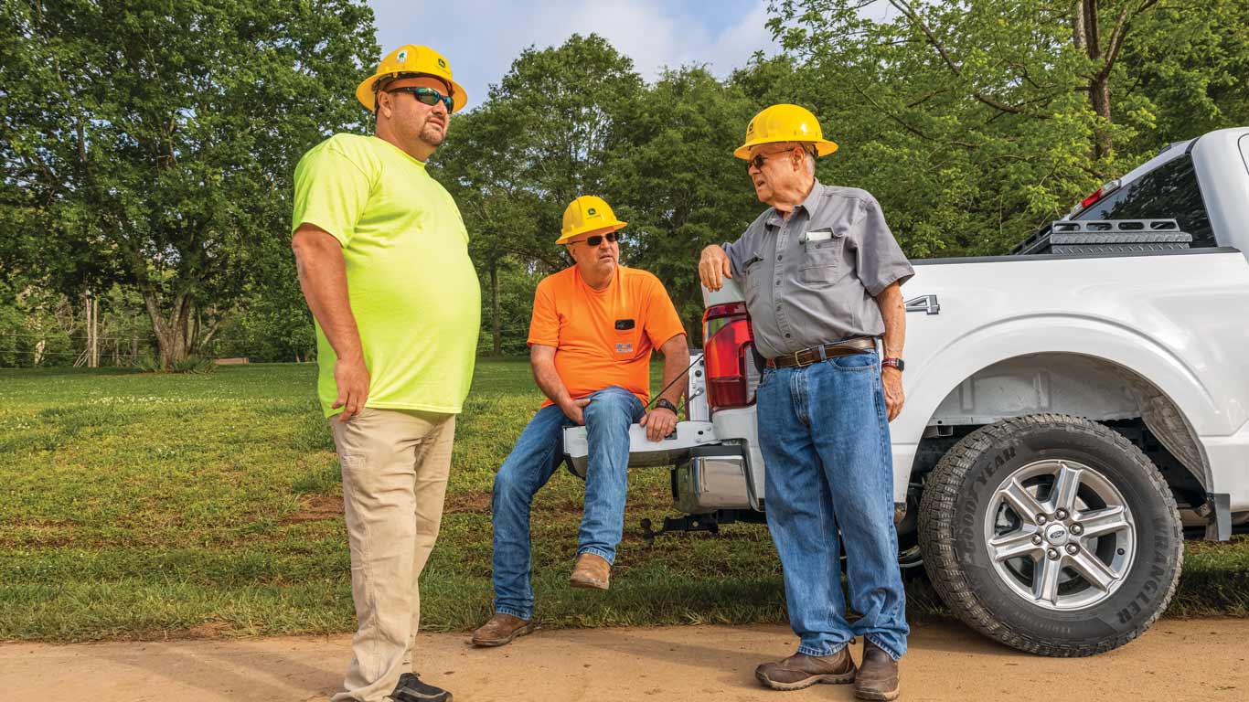 Three workers in hard hats talk near a white truck in a grassy outdoor setting.