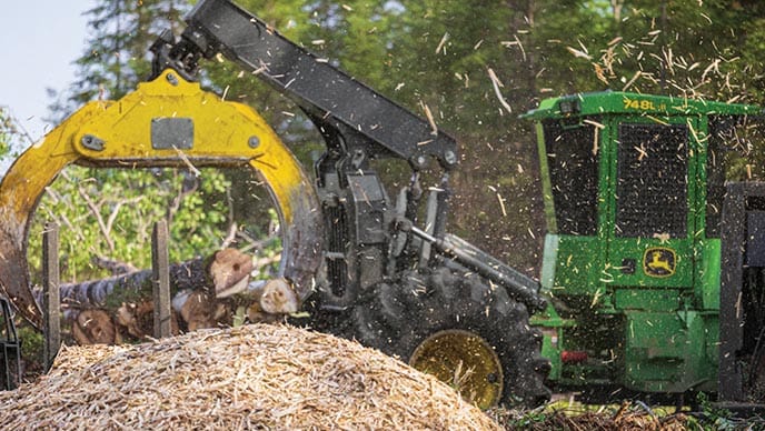 Une débardeuse à pince 748L-II de John Deere laisse tomber plusieurs arbres abattus de sa pince à côté d'une déchiqueteuse de bois qui envoie des copeaux de bois en l'air.