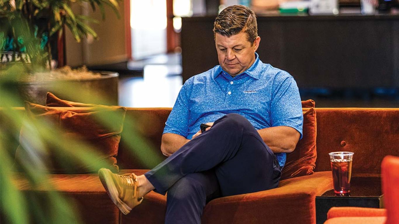 Man leaning back looking at his phone in a common area in a public space (not on a golf course)