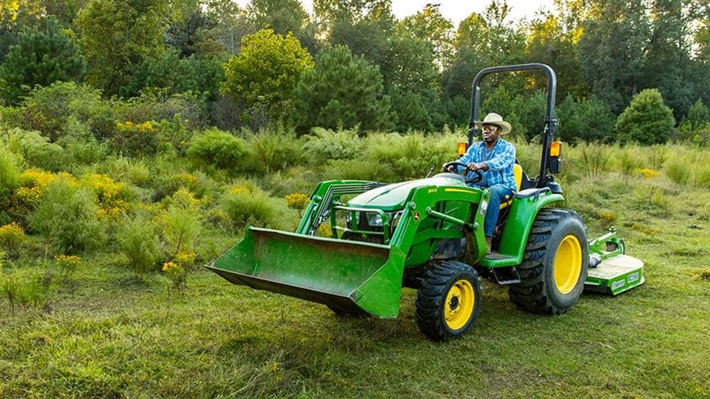 Une personne portant un chapeau de cowboy conduit un tracteur compact vert équipé d’une benne à l’avant et tractant un instrument de tonte.