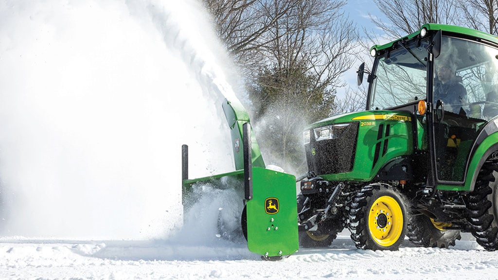 Un tracteur vert avec cabine souffle la neige à l’aide d’un instrument de déneigement.